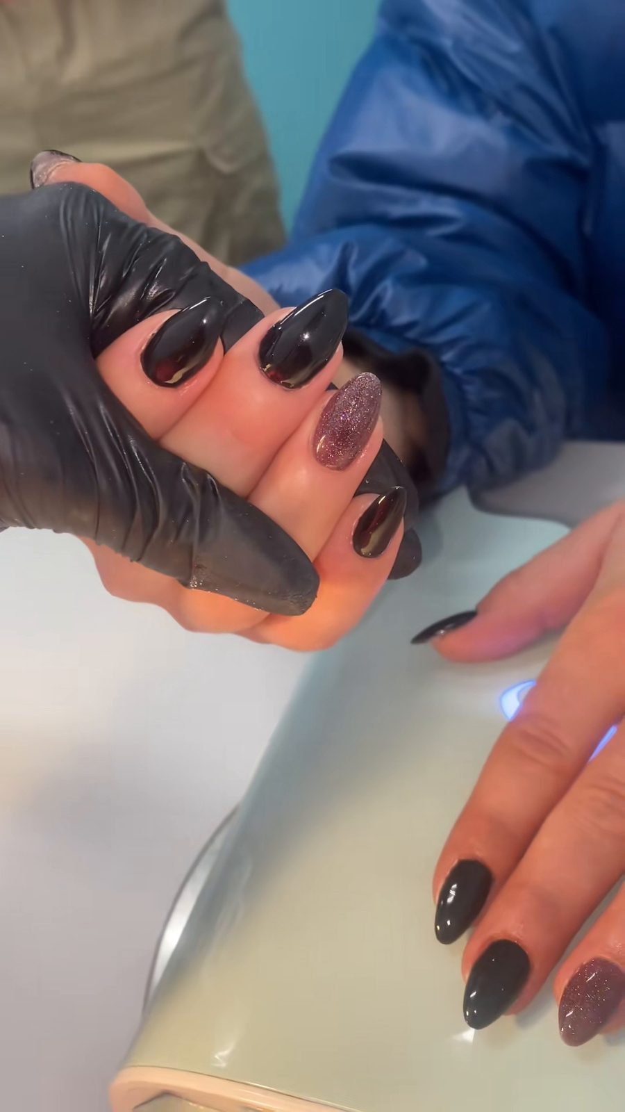 Zwarte amandelvormige nagels met één glitternagel, tegen de tealkleurige muur